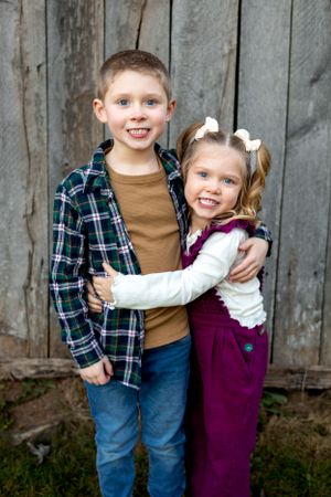 Two children in casual clothes hug and smile in front of rustic wooden wall.
