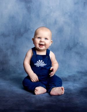 Baby in blue dinosaur overalls sitting against blue backdrop.