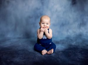 Baby in navy overalls sitting against blue background.