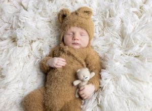 A sleeping baby wearing a brown bear outfit lies on white fluffy fabric with a small plush toy.