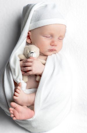 A sleeping newborn wrapped in white holds a small teddy bear.