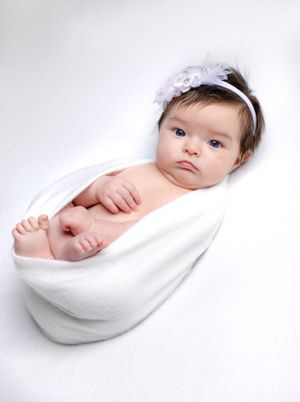 Baby wearing white headband wrapped in white blanket against light background.
