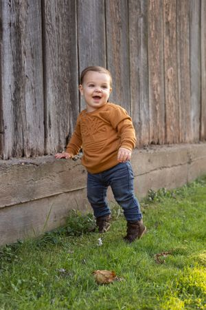 Toddler in mustard sweater plays joyfully by rustic wooden fence.