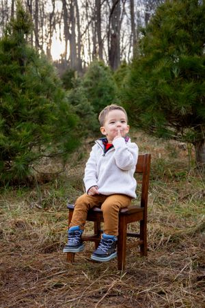 A young child sits thoughtfully on a wooden chair among evergreen trees wearing formal attire.