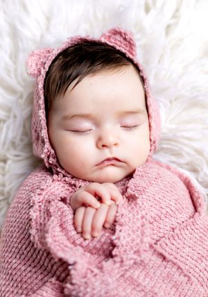 A sleeping infant wearing a pink knitted bonnet and sweater lies peacefully on white bedding.