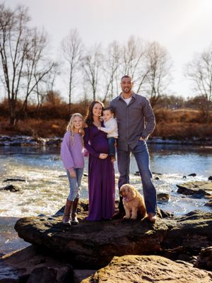 Family with dog poses on rocky riverbank during autumn portrait session.