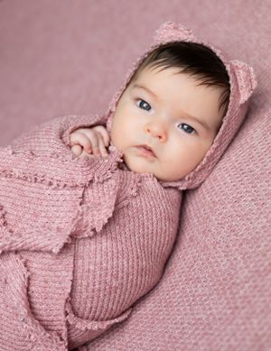 A baby wearing a pink knitted outfit gazes at camera with bright blue eyes against a soft pink background.