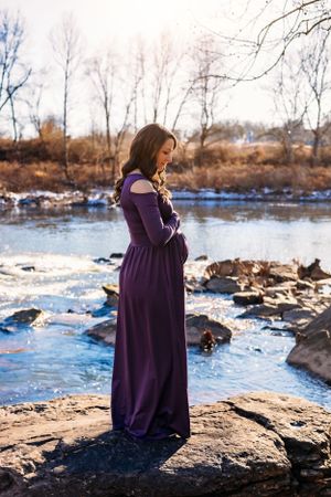 Woman in purple dress stands on rocky shore overlooking frozen river.