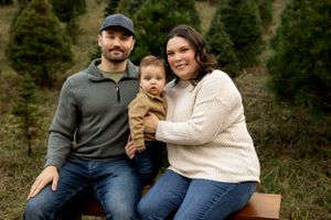 Family of three poses together on wooden bench against backdrop of evergreen trees.