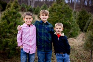 Three boys in button-down shirts standing together among evergreen trees.