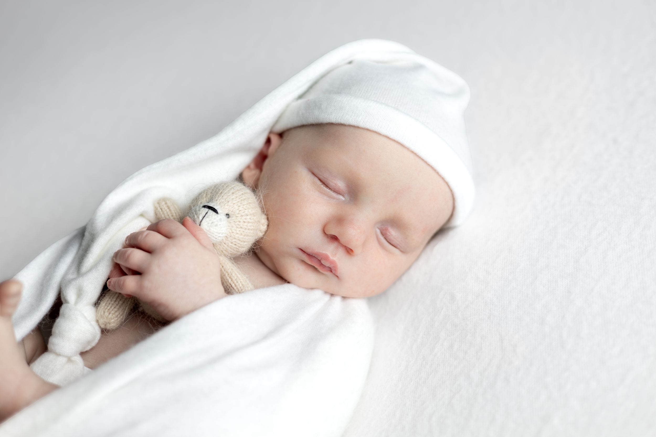 Sleeping newborn in white swaddle clutching small stuffed animal.