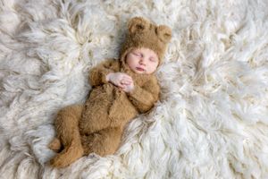 Newborn wearing cozy brown bear costume lying on white fluffy rug.