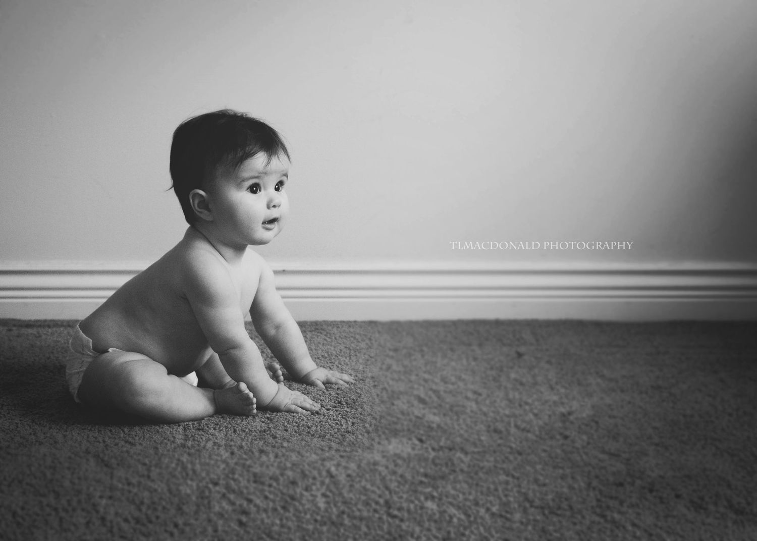 Black and white portrait of a baby in diapers sitting on carpeted floor against a wall.