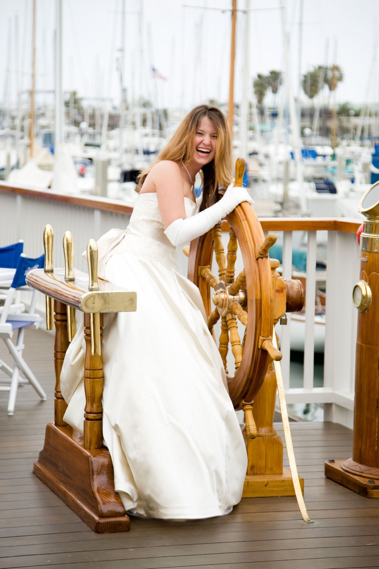 Person in white wedding dress laughs while posing at ship's wheel on marina deck.