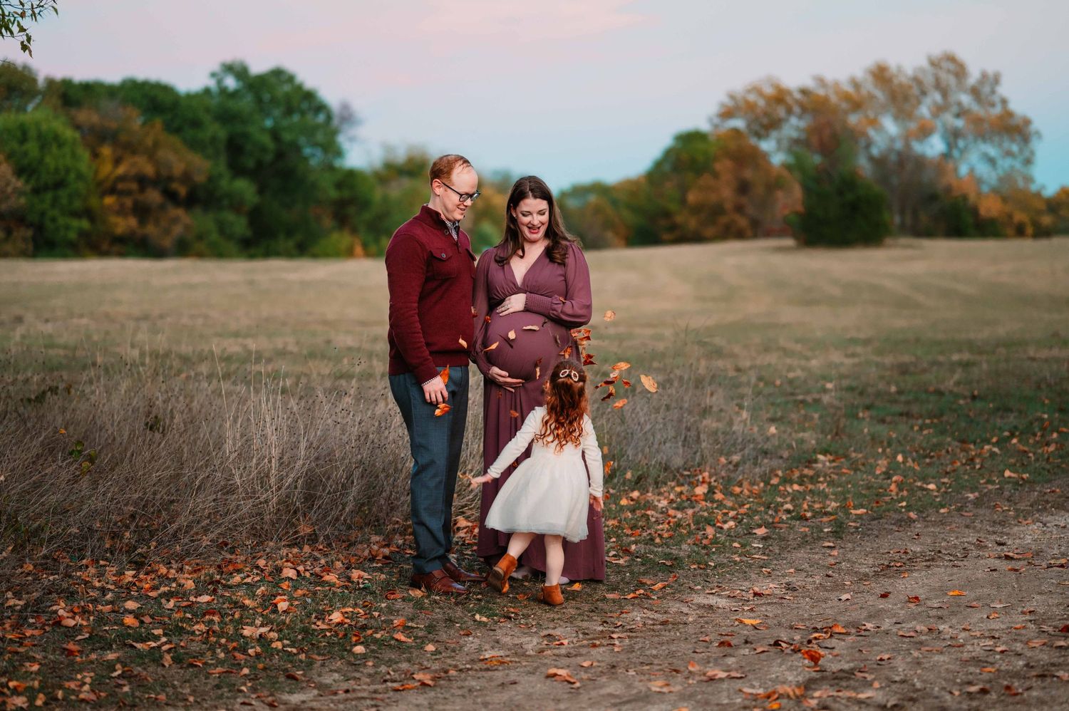 preschool aged daughter throws leaves up at parents during fall outdoor maternity photoshoot in dallas, tx