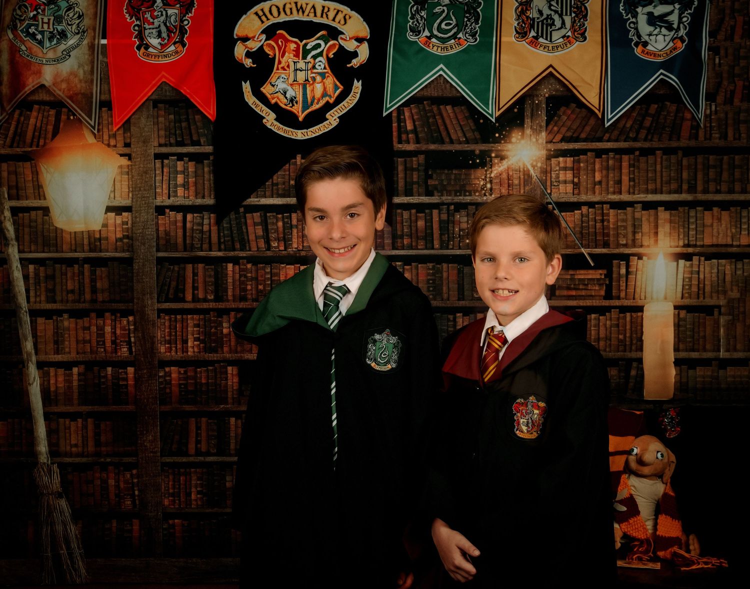 Two students pose in Hogwarts house robes against a library backdrop with magical school banners.