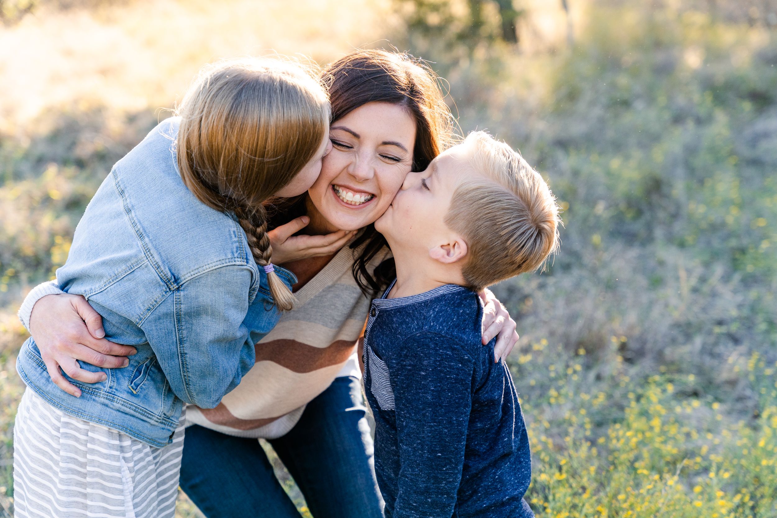 Family Session in Fort Worth, Texas. Mom kneels and smiles with her eyes closed while her two kids kiss both her cheeks.