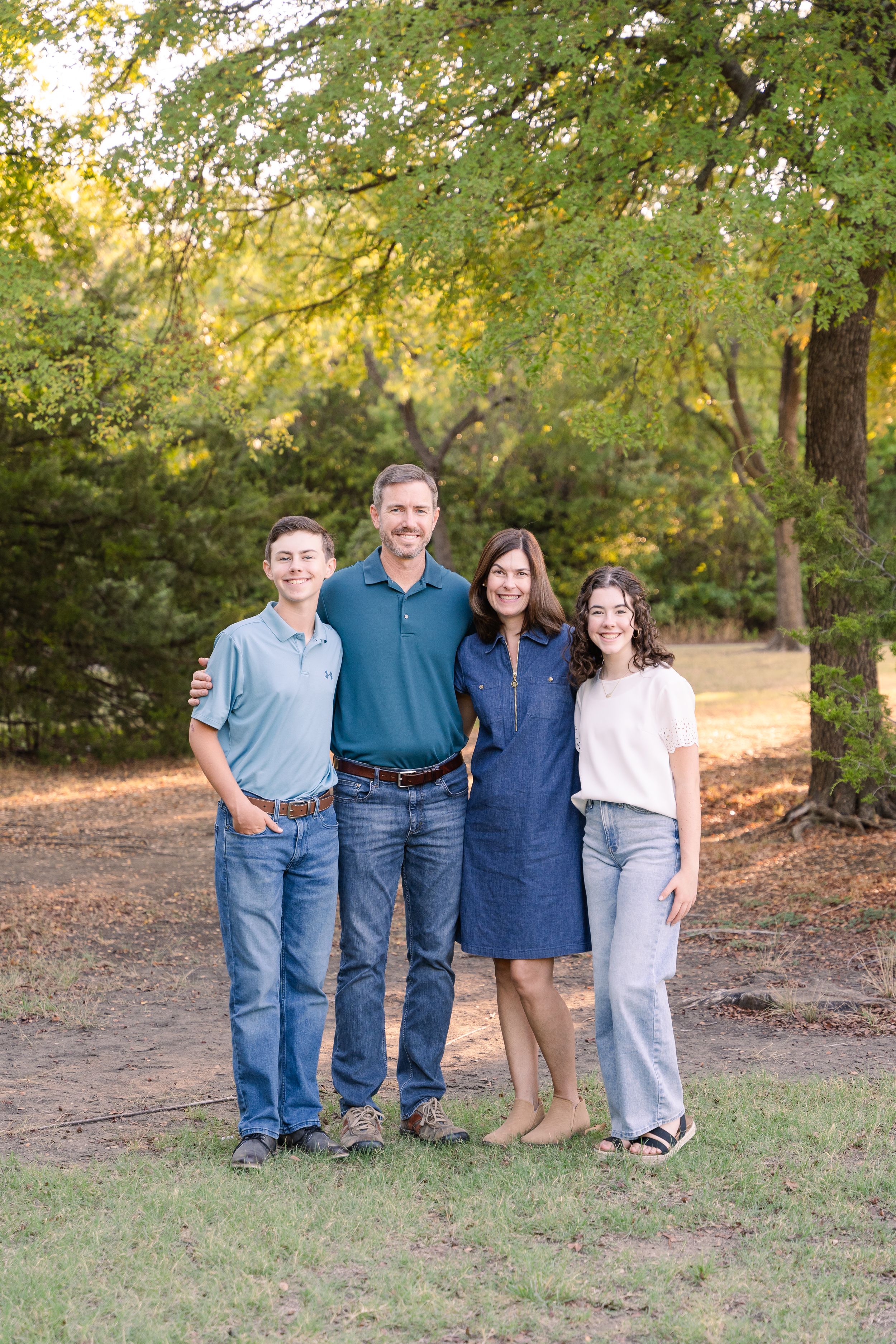Family Session in DFW, Texas. Parents with their two teenagers smile with lots of green trees behind them.