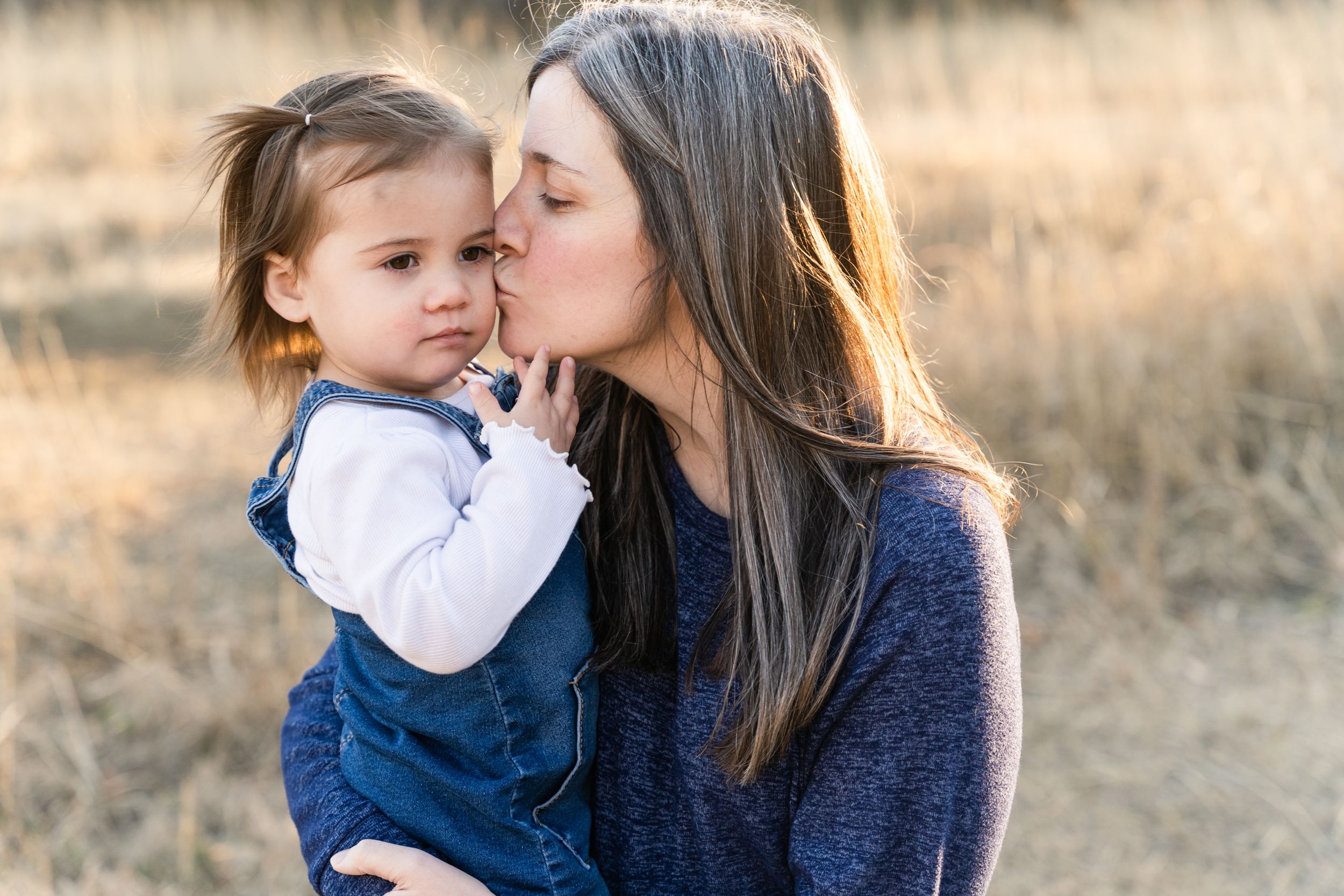 Mom holds her daughter in a golden field and kisses her cheek.