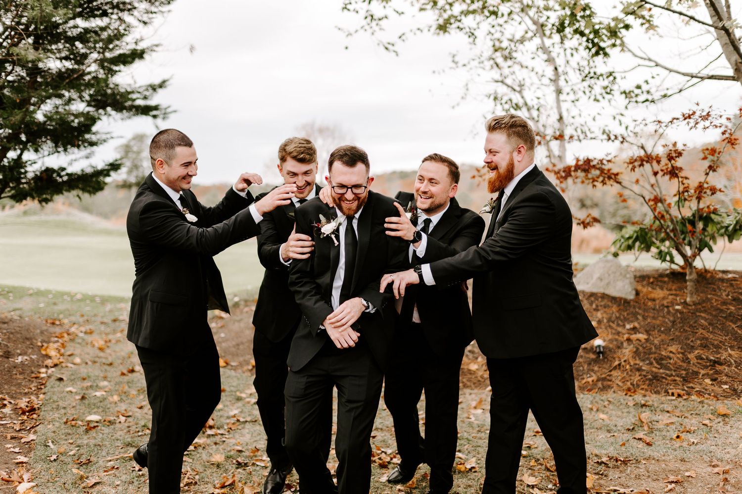 Group of groomsmen in formal attire enjoying candid moments at autumn outdoor wedding.