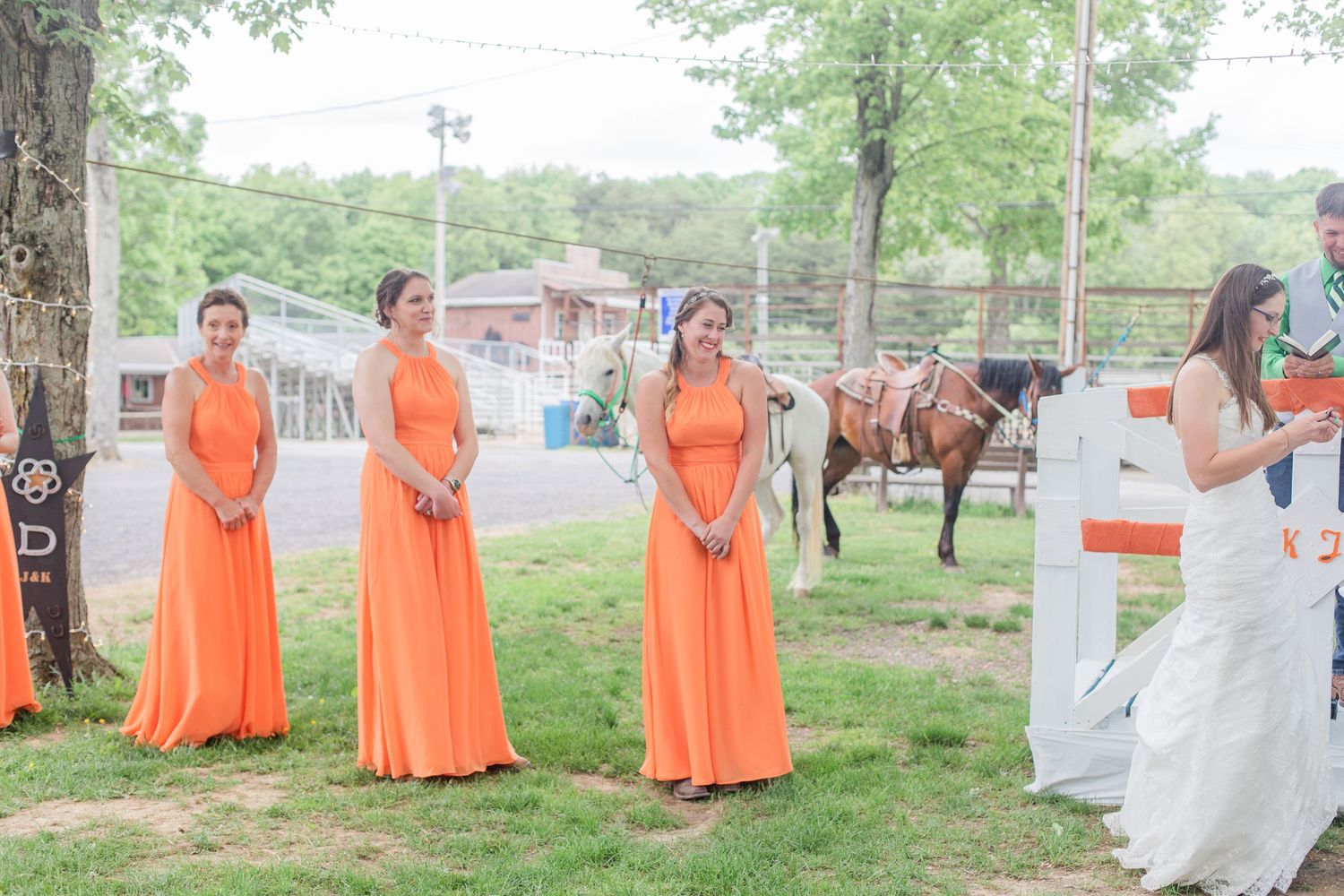 Wedding party in orange bridesmaid dresses and white gown pose together in a grassy outdoor setting.
