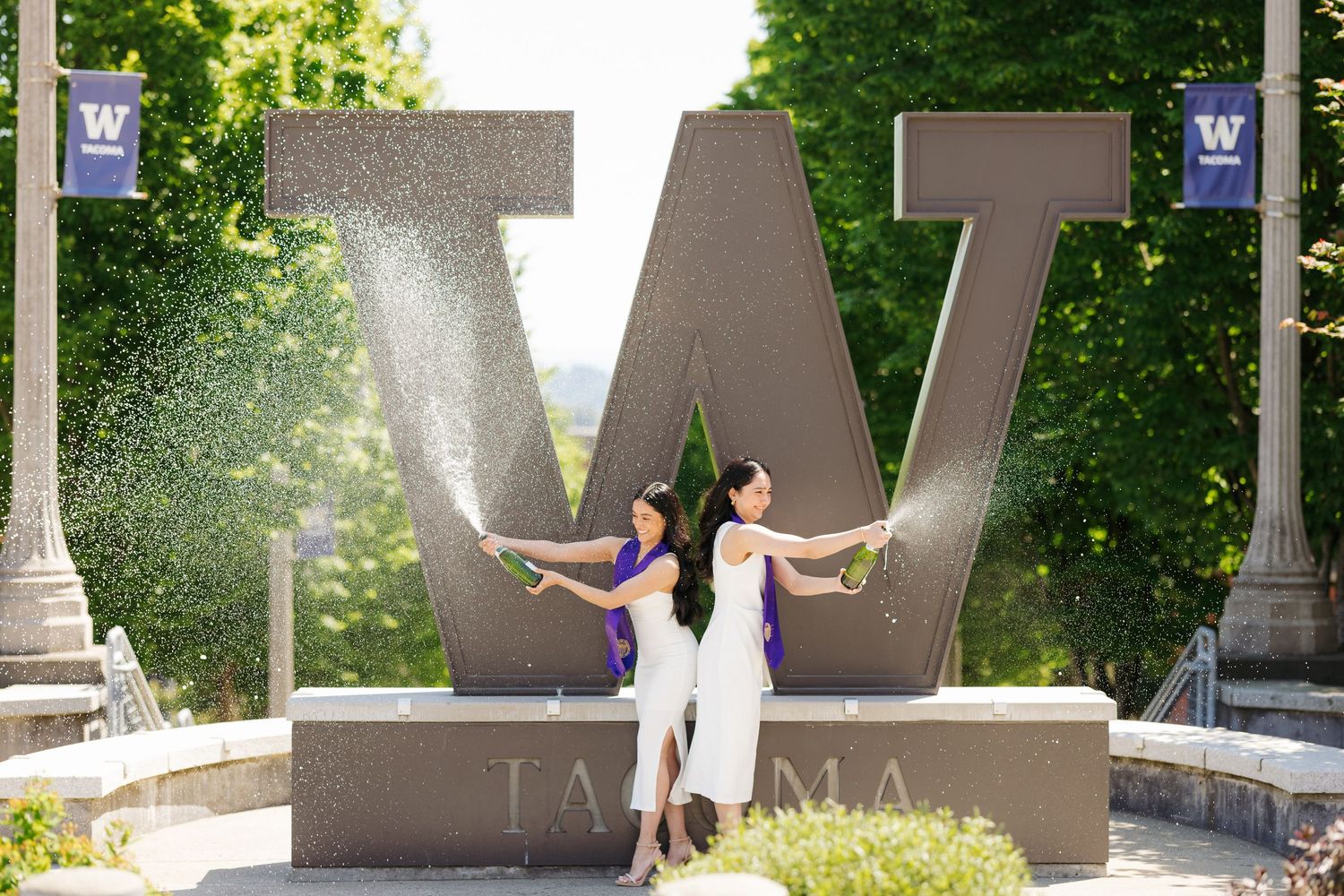 Two graduates celebrating by a large W monument with water spraying in background.