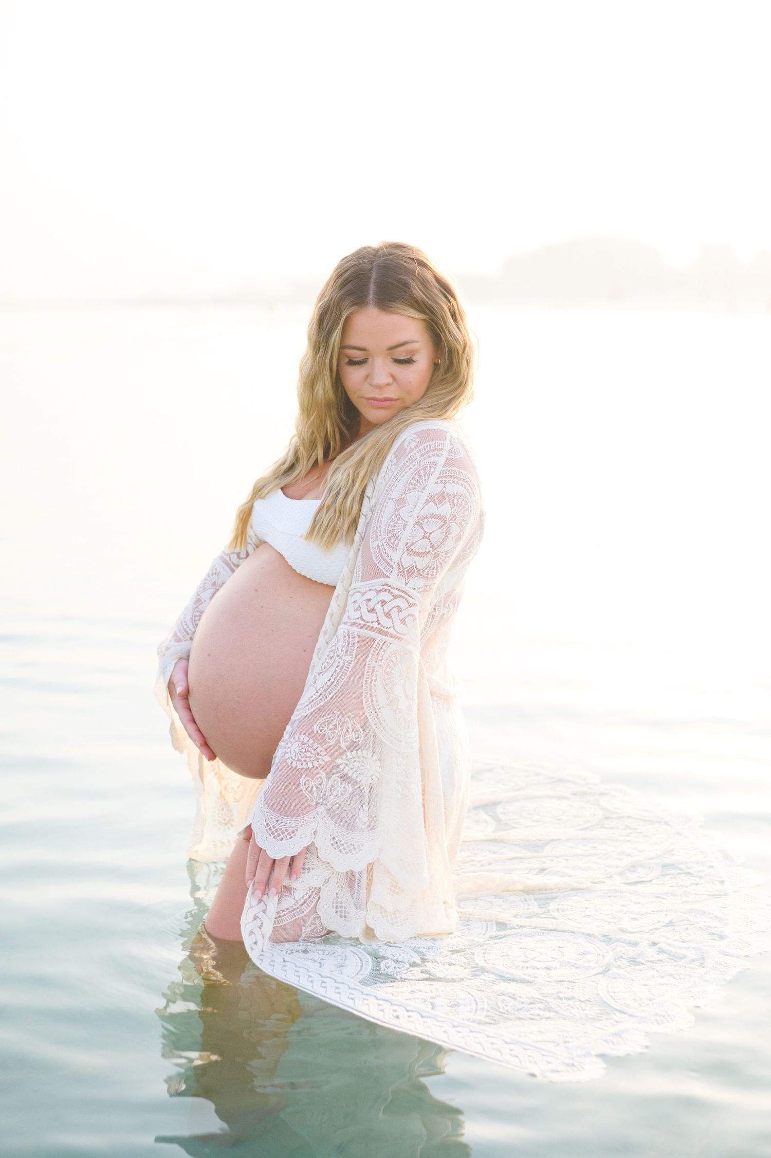 Ethereal maternity photo of pregnant woman in white lace dress standing in water at sunset.