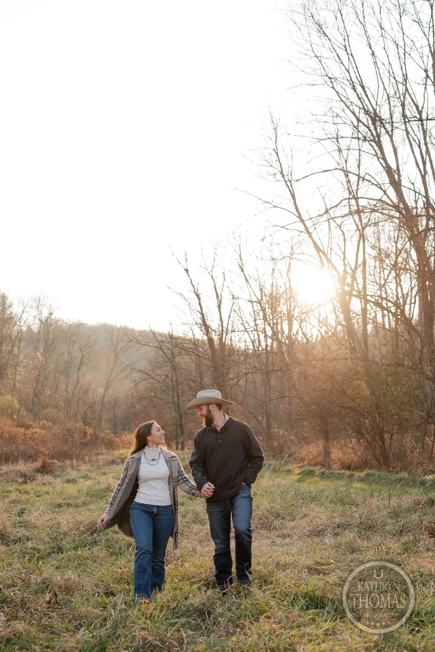 A couple walks hand in hand through a rustic field during sunset with leafless trees behind them.