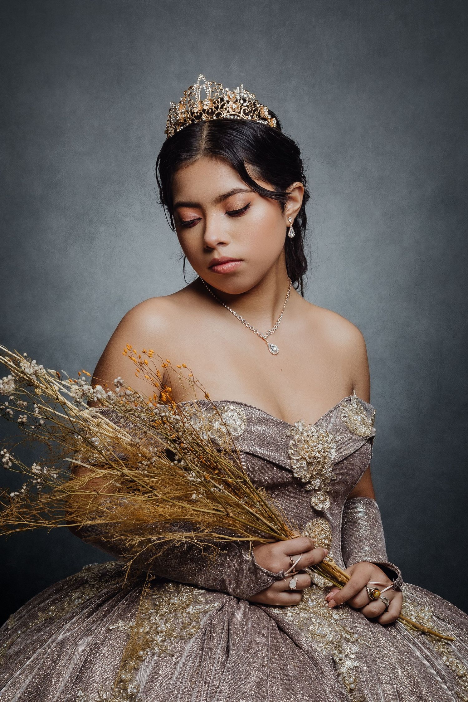 Regal portrait of woman in sparkly gown and crown holding dried flowers against grey background.