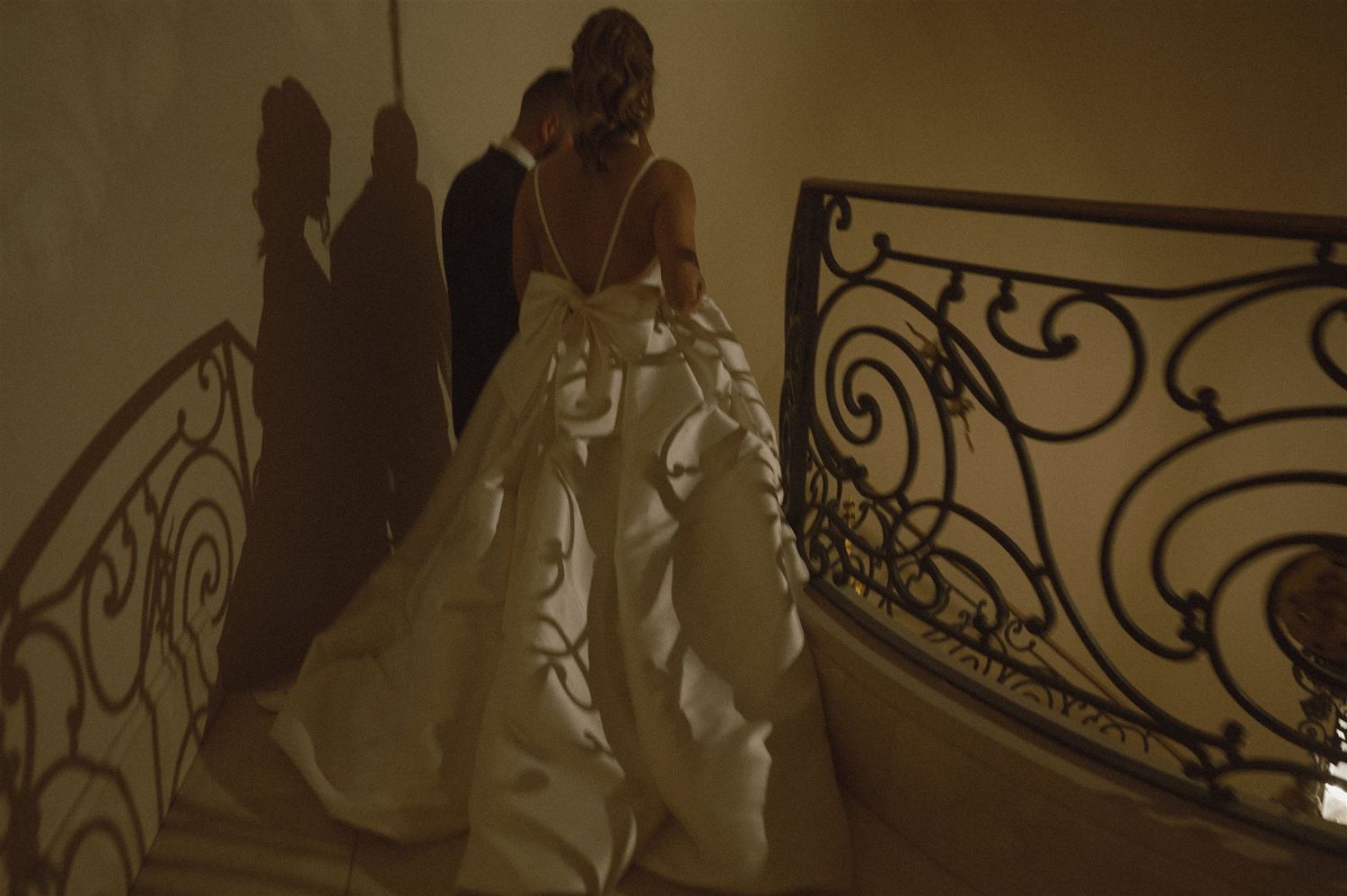 A bride in a flowing white wedding gown stands on a staircase as dramatic shadows reflect on the wall from ornate railings.