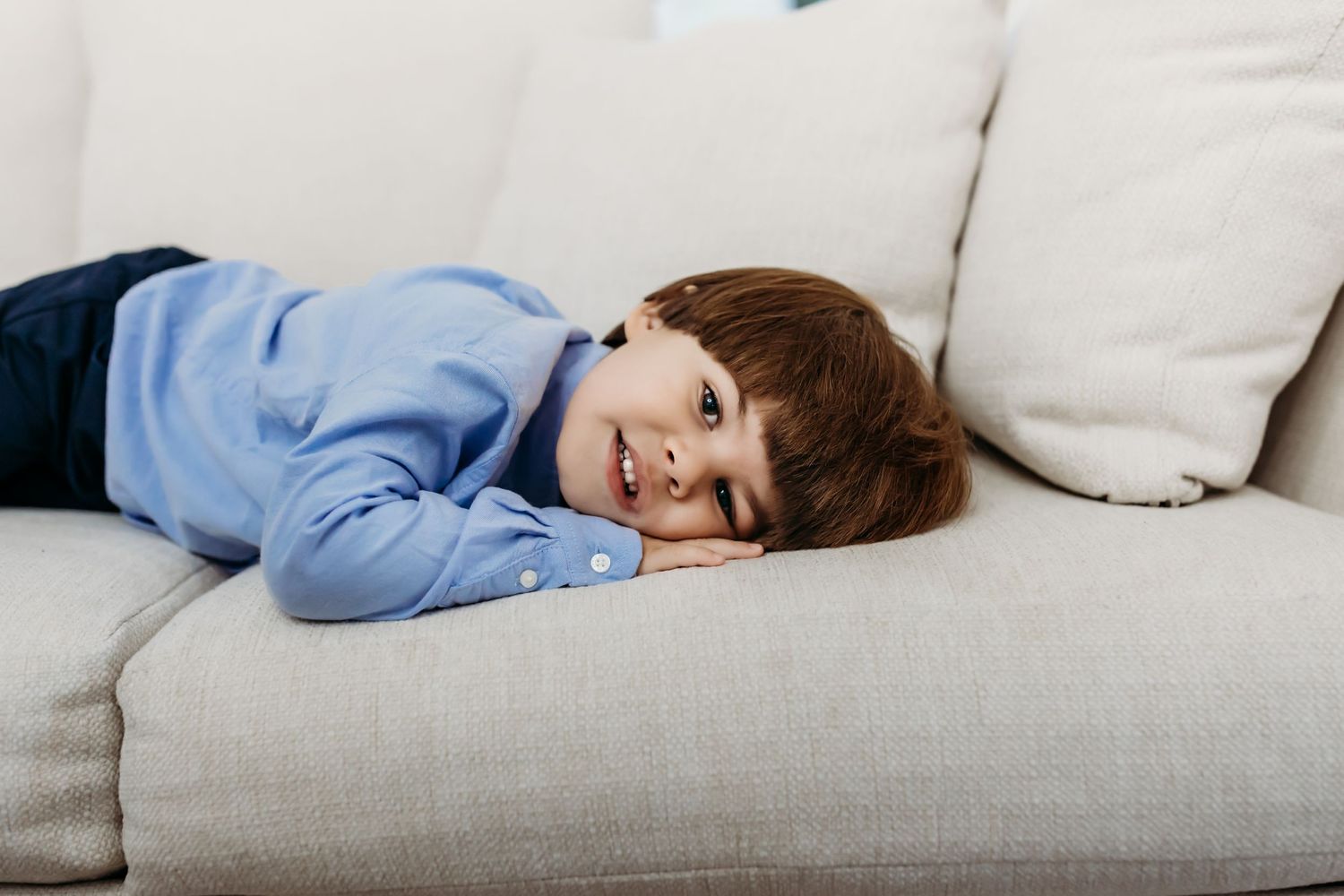 Child in blue sweater relaxing on gray couch with white pillows.