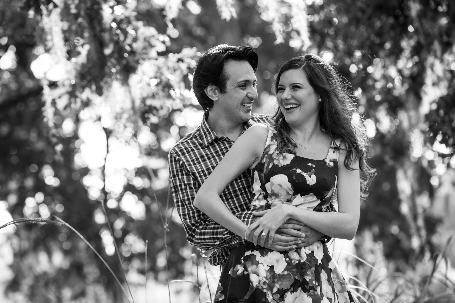 A couple in black and white laughs together while holding their dog in a wooded outdoor setting.