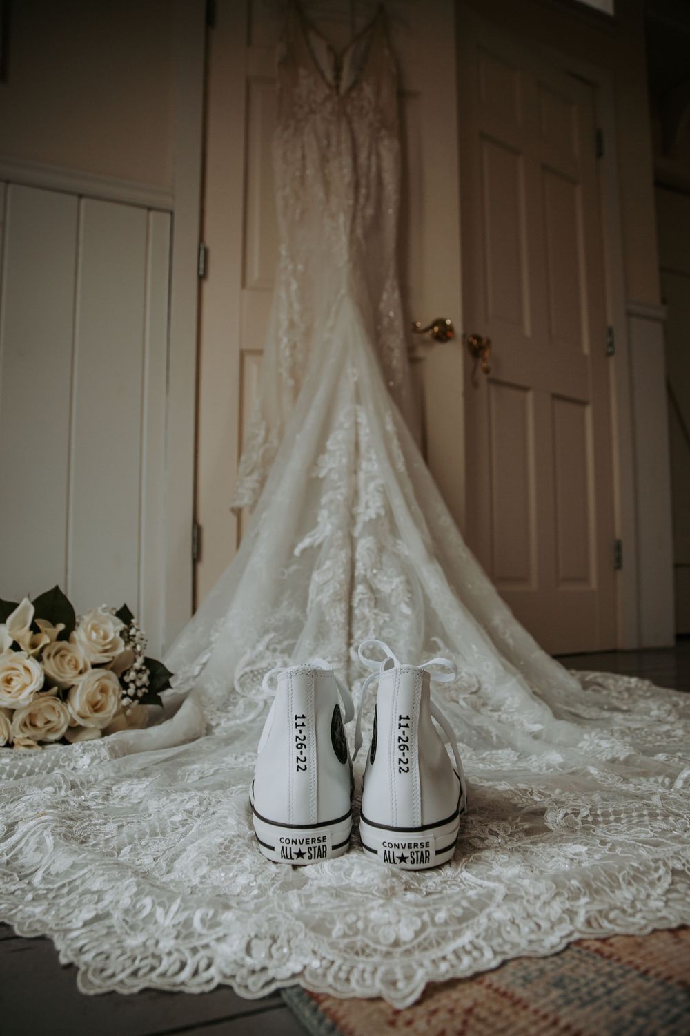 White Converse wedding sneakers sit before a lace wedding gown hanging on a door, next to a bouquet of cream roses.