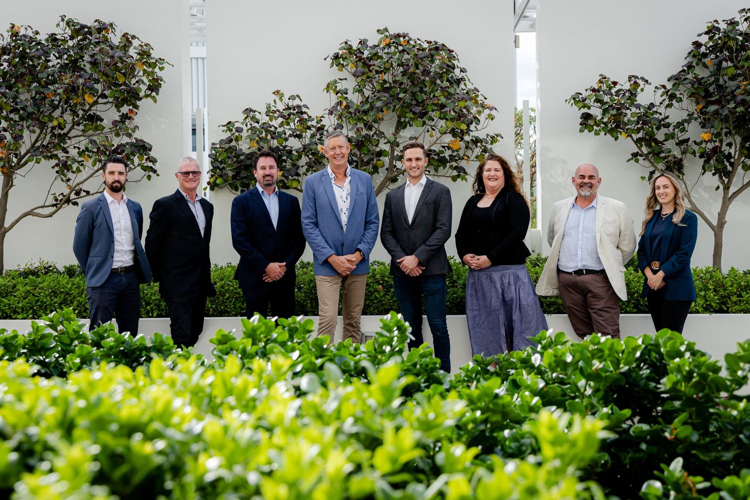 Professional business team standing together in front of green landscaping in an outdoor setting.