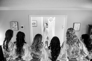 Bridesmaids in robes admire wedding dress hanging in doorway, black and white.