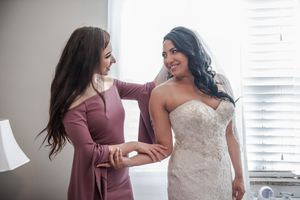 Bridesmaid in pink dress helping smiling bride with her lace wedding gown before the ceremony.