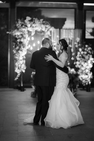 Bride and groom share first dance in front of floral arch with twinkling lights in black and white.