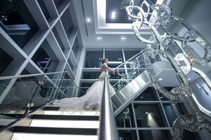 Bride in flowing white gown poses on grand staircase beneath dramatic crystal chandelier.