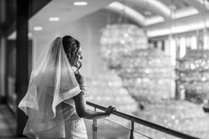 Elegant bride in lace gown and veil gazes over balcony railing in black and white wedding portrait.