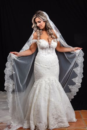 Blonde bride in off-shoulder lace mermaid gown displays cathedral veil with lace trim.