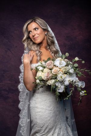 Bride in lace mermaid gown holds white and blush bouquet wearing cathedral veil in studio portrait.