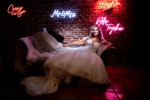 Bride in white gown poses on vintage sofa surrounded by romantic neon signs reading Mr & Mrs at wedding venue.