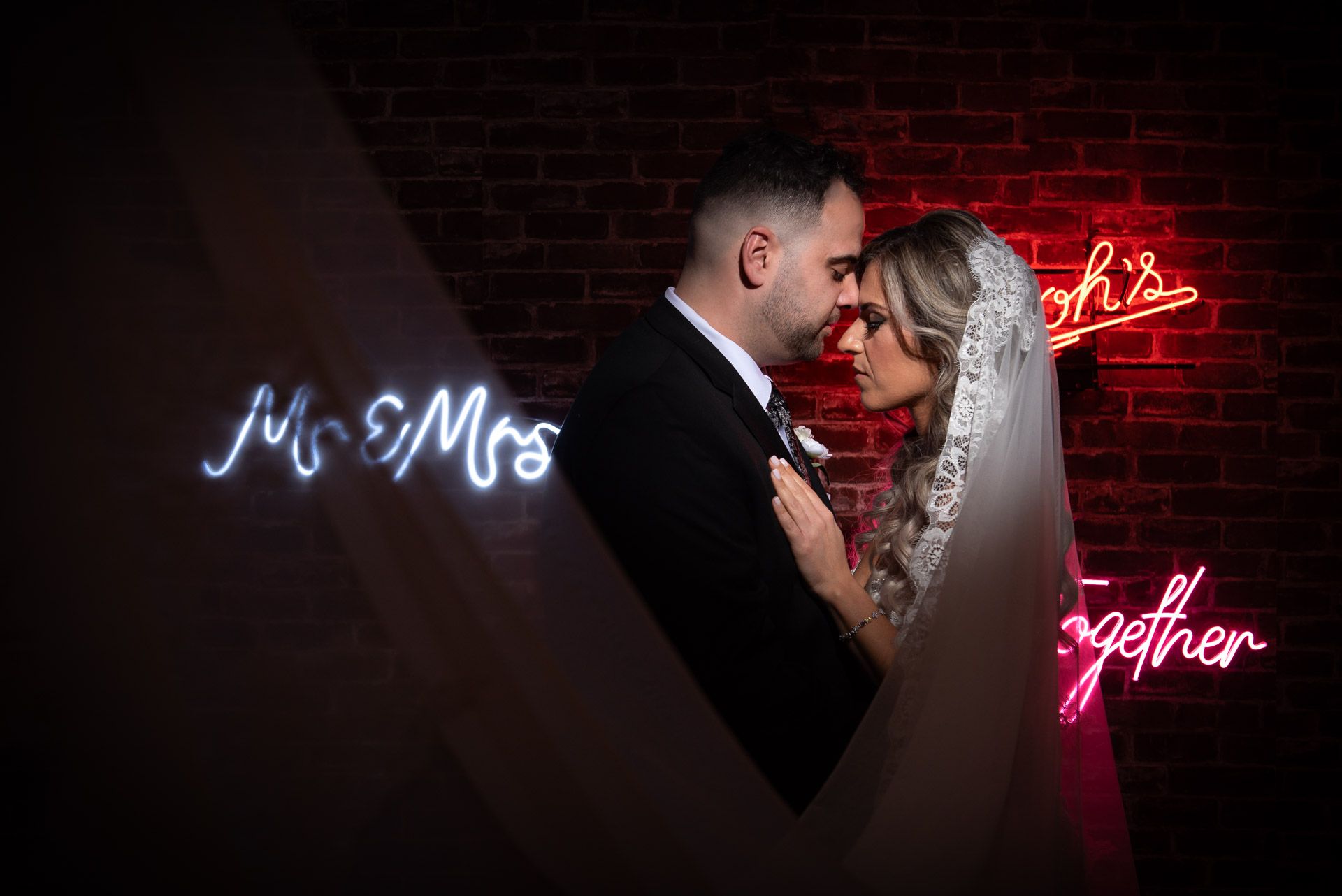 Bride and groom share intimate moment in front of glowing Mr & Mrs neon sign on brick wall.