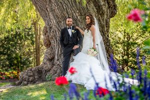 Elegant bride and groom posing beside a large tree surrounded by colorful garden flowers.