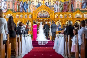 Bride in white gown walking down red-carpeted aisle of ornate Orthodox church during ceremony.