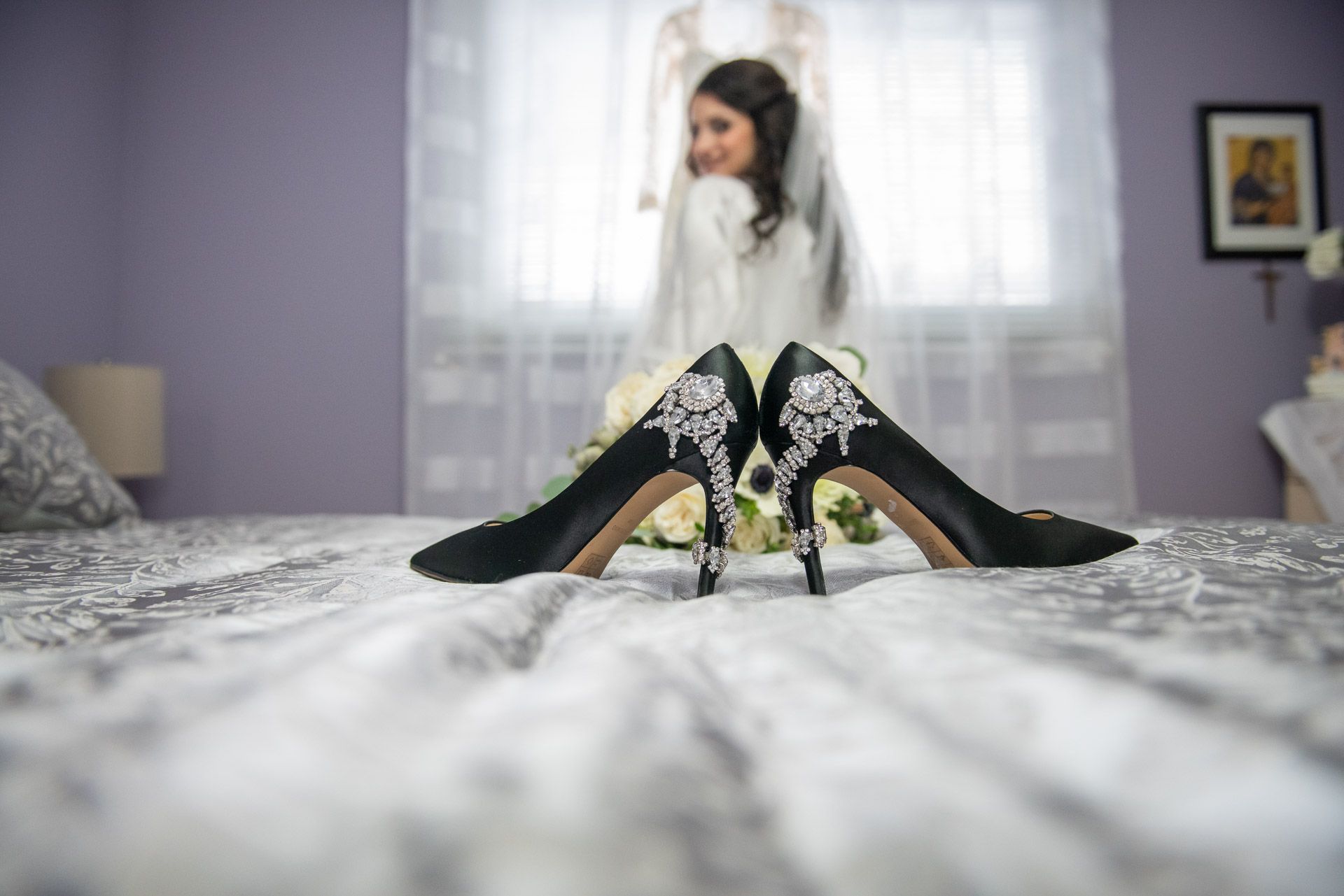 Elegant black bridal heels with jeweled detail displayed on bed with bride in background.