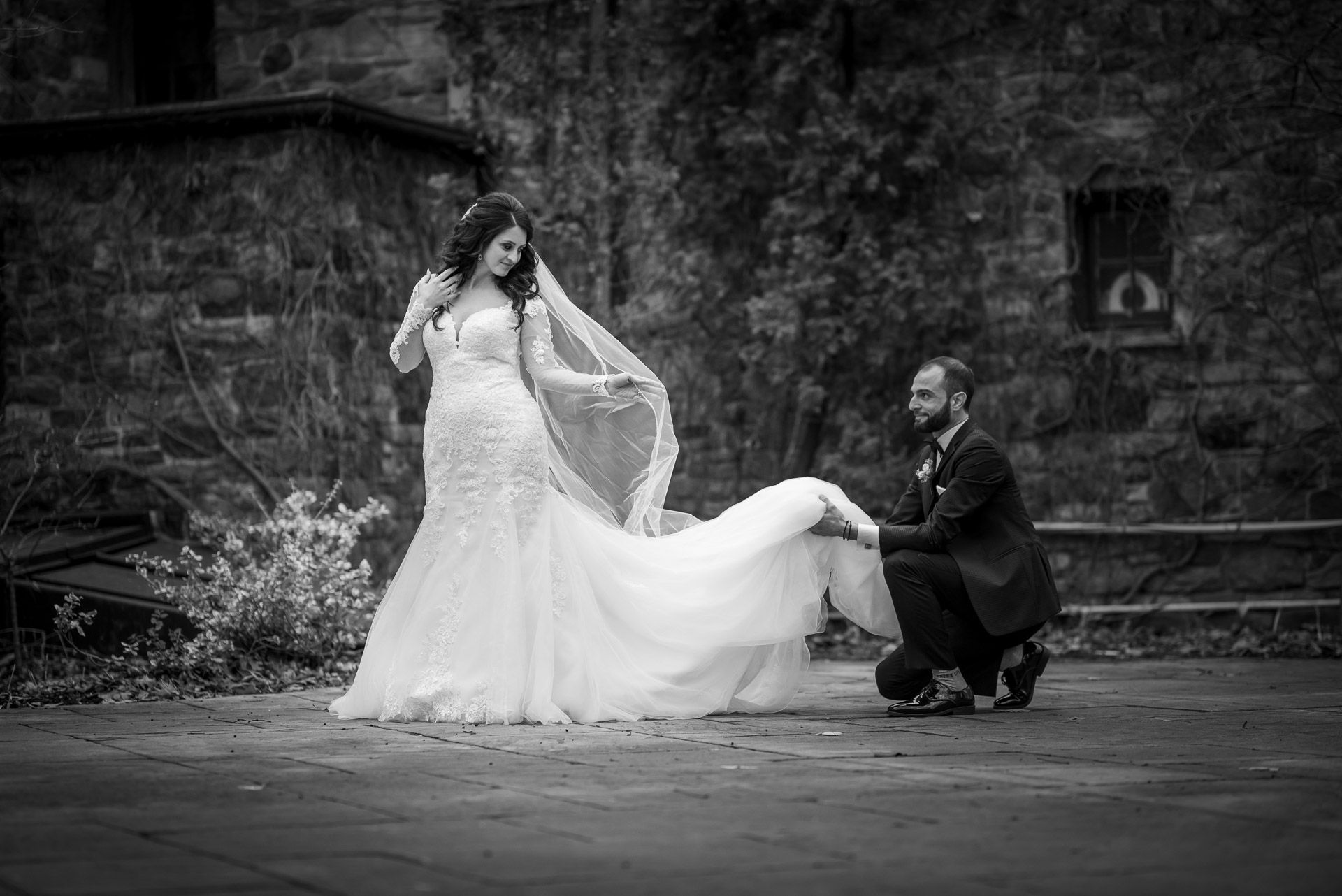 Groom kneels to arrange bride's wedding gown train in classic black and white outdoor portrait.