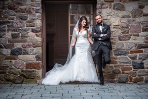 Bride and groom pose confidently against rustic stone wall in elegant wedding attire.