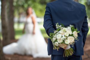 Groom in navy suit hides white floral bouquet behind back as bride stands in distance.