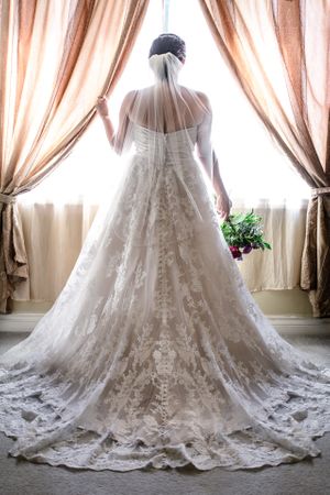 Bride in elegant lace wedding gown with veil stands before a window holding a bouquet.